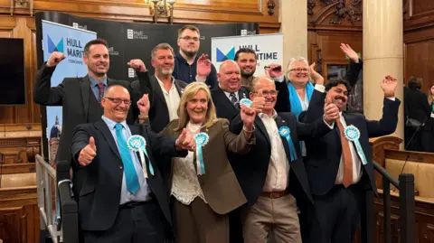 A group of women and men dressed in suits, wearing blue and white badges, with their arms in the air in celebration. 
