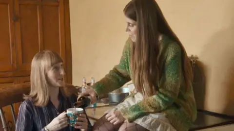 Gemma pours Mildred some red wine in a mug