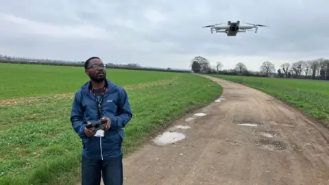 Dr Emmanuel Zuza stands on the track in the middle of a young wheat field and uses a console to fly a white drone that hovers just above head height
