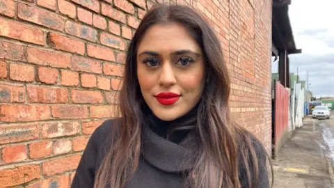 A woman with long brown hair and wearing red lipstick is looking at the camera. She is wearing a grey top and standing outside by a brick wall.