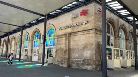 An exterior view of Hull Paragon Interchange railway station, with a stone facade featuring large arched windows filled with blue and green glass panels. The words “Hull Paragon Interchange” and the red British Rail logo are mounted on the wall. A modern canopy supported by dark columns covers the pavement in front.