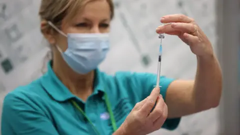 A nurse in uniform and wearing a face mas is drawing vaccine from a vial with a needle