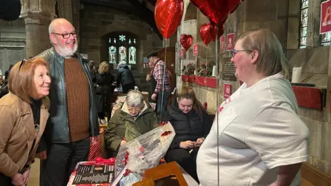 Jackie Southall and Bob Wright who are both in their 60s are smiling at Natalie Walsh who is a larger woman wearing a white top. They are in a church and there are lots of people behind and red balloons tied to the windows