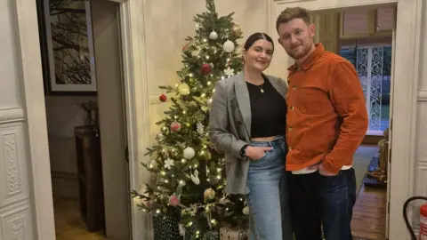 Laura O'Reily A woman in a blazer and black jacket and jeans stands in front of a Christmas tree with a man in an orange shirt and jeans. They are in a hallway with cream ornate wall panelling and other rooms can be seen beyond.