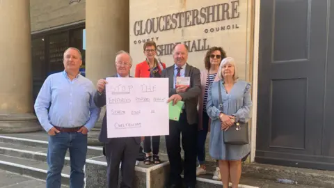 A group of residents outside Gloucestershire County Council Shire Hall with a placard demanding a stop to the parking changes.