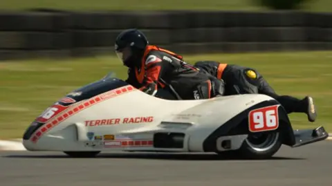 A white sidecar, with red writing reading 'Terrier Racing' on the side, and the number 96 printed above the wheel arch. The driver is wearing black and red leathers, a black helmet, and an orange hi-vis vest. To the right, you can see the passenger leaning over the right hand side of the sidecar to counterbalance it. She's also wearing black leathers, a black helmet, and an orange hi-vis vest. The sidecar and the racers are in focus, but the background is blurred. 