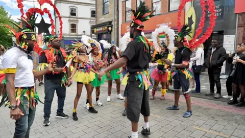 Stephen Huntley/BBC A group of dancers wearing costumes are stood together on a street in Luton