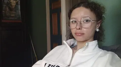 Orianah Upland A young woman has brown curly hair tied back in a short ponytail, she is wearing large gold frame glasses and a white hoodie. She is sitting in a room with dark green walls