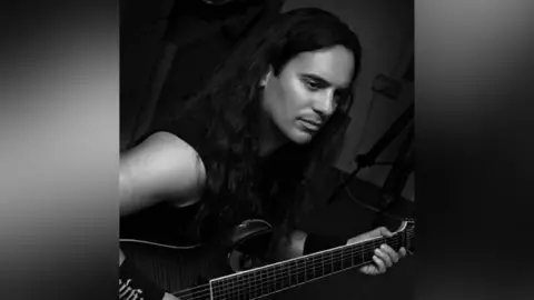 Ruth Hepner Black-and-white photo of Dan. He has long dark hair and is playing a guitar while looking at something out of shot.