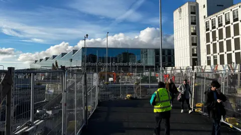 A picture of Belfast Grand Central Station, a large glass transport hub, from the view of a walkway which goes through ongoing building work.