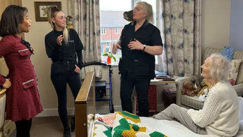 Kimberley Piper/BBC An elderly care home resident is sat on a bed and looking at the two handlers from Amazing Animals. Both the handlers each have a monkey on their shoulder.