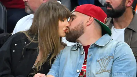 Getty Images Taylor Swift and Travis Kelce kiss during a game between the New York Knicks and the Cleveland Cavaliers in Cleveland, Ohio, earlier this year. She is wearing a black jacket, he is wearing a blue deim shirt and red baseball cap.