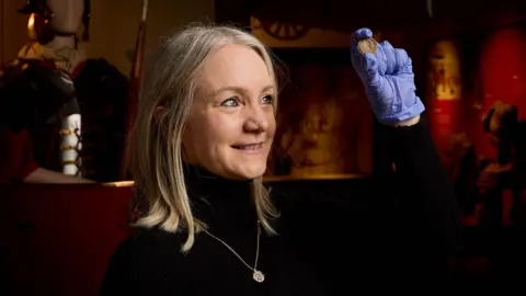 Wakefield Council A woman with blonde hair wearing a black jumper and a gold medallion necklace, and blue gloved holds a coin-shaped white disc.