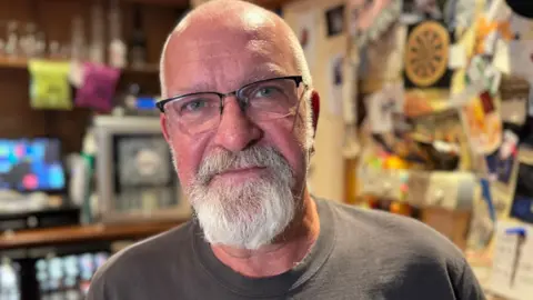 A man with glasses and a beard stands indoors with shelves and photos visible in the background.
