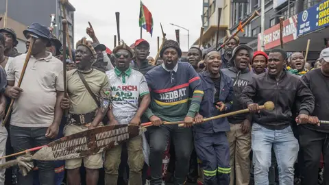 AFP via Getty Images Members of the Zulu regiment known as the 'Amabutho' holding sticks march during a demonstration 
