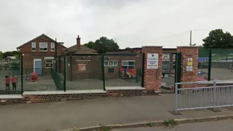 Google Maps A street view image of the school, a red-brick building with a concrete front yard. The building is surrounded by tall, green fences and features a sign at the entrance pillar.