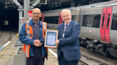 A man in an orange hi-viz jacket shakes the hand of another man holding a certificate which says 'Congratulations on your long service'. The two men smile at the camera as they stand on a train platform, with a silver and red train visible on the track to the side of them.