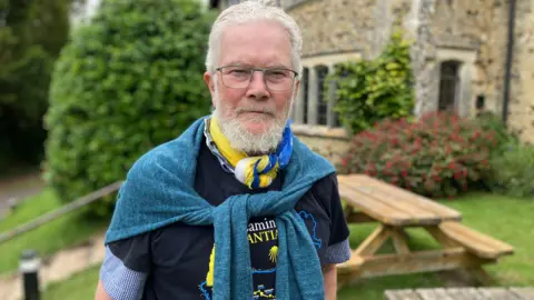 Victor Ient with glasses and a grey beard standing in a garden, wearing a scarf and sweater over the shoulders, with a picnic table and stone building in the background.
