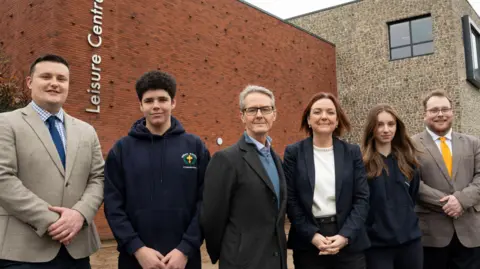 A group of six people standing in a row, looking towards the camera and smiling. Behind them is a red brick building that has a sign that says leisure centre