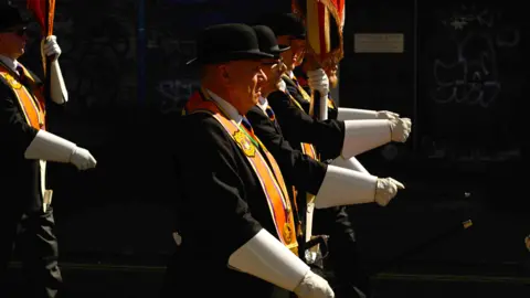 PA Media A number of men dressed in black with white gloves march on the street in Belfast. They are wearing Orange sashes and black bowler hats.