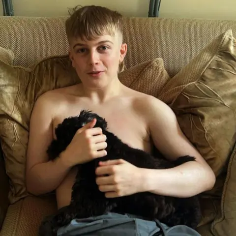Martell Blackford Teenager Jacob Holman. He is sitting on a sofa. He has no t-shirt on and is wearing grey shorts. He is cuddling a dog and smiling at the camera. 