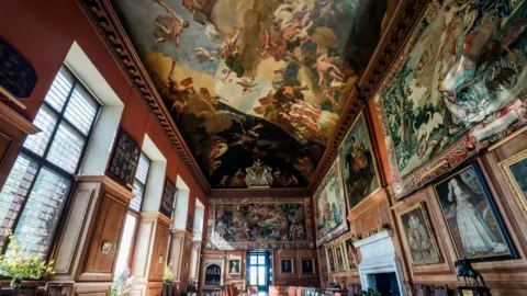 Tony Marsh The inside of a stately home, showing lots of work of art on the walls and a painted ceiling. There are windows to the left, a doorway at the end and wooden panelled walls. Tapestries are also on the walls. 