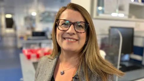 A woman is standing in a lab and smiling to the camera. She's wearing glasses and a suit jacket. She has a necklace with a heart pendant on a silver chain. She has long brown hair. 