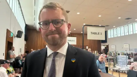 A man with short blonde hair and a short blonde beard smiles tightly at the camera in a room with people and tables.