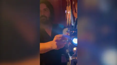 A front shot of a man operating an old school film camera. He is looking off at the screen as the film runs