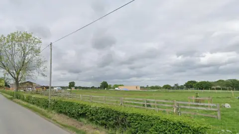 Google A view of a field from a country lane. There is a line of trees in the distance and, to the left of the shot in the background, the dairy farm near to which the zoo would be built. 