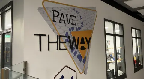 A yellow triangle is on a white wall with the words 'the way' written in black capital letters. The letter A in the word Way is stylised into an arrow logo.