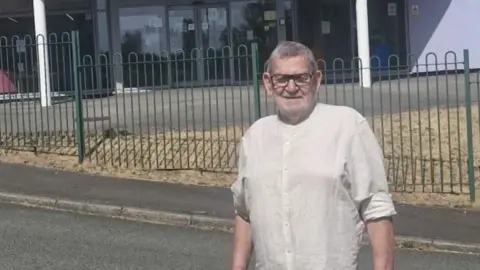 An man with grey hair, wearing glasses and a white shirt, outside an education building. There's also dry yellow grass and big green gates behind him. 