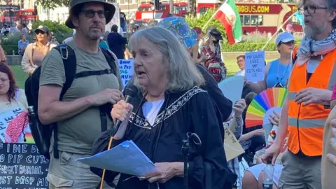 Joy Dove speaking at the rally on Parliament Square. She has shoulder-length grey hair and is holding a microphone in one hand and her speech in the other. A sign language interpreter stands to her left. She is surrounded by protestors holding placards.