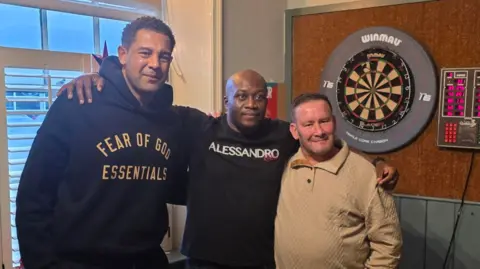 Three people in a pub, with a dart board behind them, with the middle man wrapping his arms around the two men on either side of him.