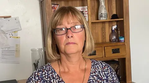 Kate Bradbrook/BBC Wendy Randall pictured alone wearing glasses and a long sleeved top. She is also wearing a silver necklace and is standing in a home office environment.
