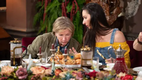 BBC/Studio Lambert/Euan Cherry A blonde woman and a younger dark-haired woman sat around a table covered in food deep in discussion - Amanda Collier and Rachel Duffy in The Traitors