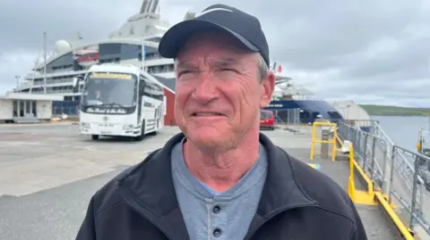 Rich Lancellot is standing outside on at aferry terminal. He is wearing a dark blue cap and a black fleece over a grey, buttoned top.