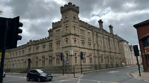 A view of the exterior of County Hall in Ipswich. The building is made of large grey bricks and has a similar look to a castle. People can be seen walking past.