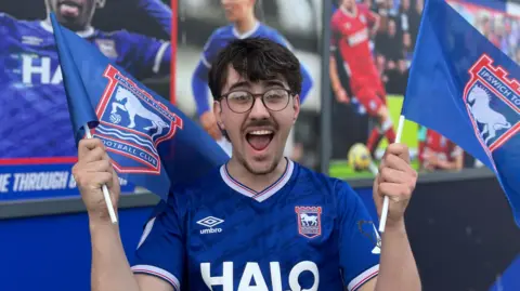 Aimee Dexter/BBC Jack Richardson is holding two flags either side of him. He has a happy expression and has short brown hair. He is wearing black framed glasses and a blue Ipswich Town FC football T-shirt.