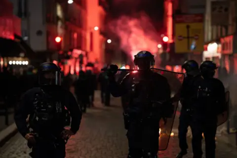 Jerome Gilles/NurPhoto/Getty Images Police use tear gas to disperse protesters at Place de la Republique in Paris, France during a demonstration on June 30, 2024. Police in riot gear stand in front as red lights and smoke billow behind them at night.