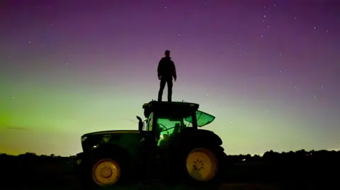 EmWin/Weather Watchers A man stands on a tractor with the Northern Lights in the background in Frieth, Buckinghamshire