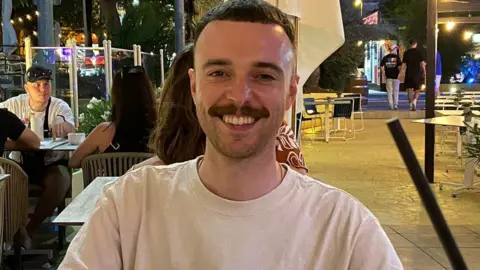 Lewis McKie All smiles on holiday, but certainly not on test day, a man grins at the camera. People in the background await their dinner.