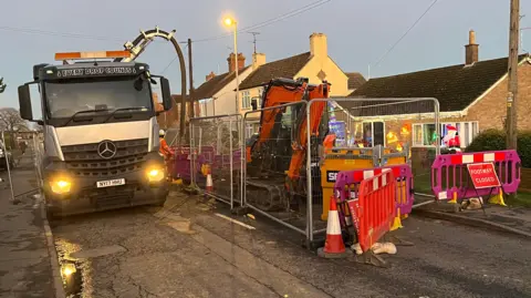 John Devine/BBC A white lorry on the road next to work equipment, including an orange digger and road diversion signs