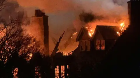 Flames engulf a large building at night, with thick smoke billowing into the sky as fire spreads through the roof and windows.