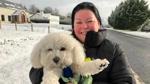 Eilena McKeever holds her poodle Buddy. She has black hair and is wearing a winter coat. In the background snow is visible on the roads and streets. A house can be seen with snow in the garden and on its roof.