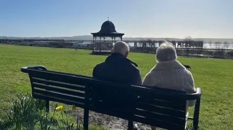 A wide grassy park on a sunny day, with two people sitting on a black bench facing away from the camera. In the distance stands an ornate bandstand and a large body of water. Daffodils bloom near the bench, and the sky is clear and bright.