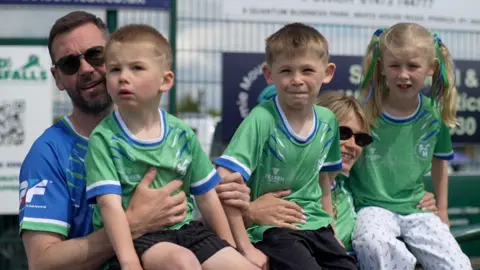 Connor Bennett/BBC A row of three children - two boys with blond and a girl with long blonde hair in bunches - sitting on a barrier, held in a hug from behind by a man with a blue football top, brown hair, beard and sunglasses to the left, and a woman with blonde hair, green football top and sunglasses between the second boy, and the girl. All three children are in green football tops.