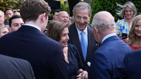 Getty Images Nancy Pelosi in a blue dress chats with the King in a pinstripe suit at the garden party in the gardens of the British Embassy in Washington. Other guests watch on in the background. 