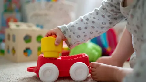 A baby wearing a grey leopard print baby grow plays with toys.