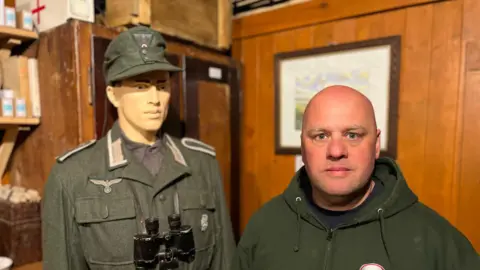 Malcolm Amy, on the right, wearing a green hoodie. He is with a mannequin in army uniform and binoculars. Behind him is a wooden wall and a painting.